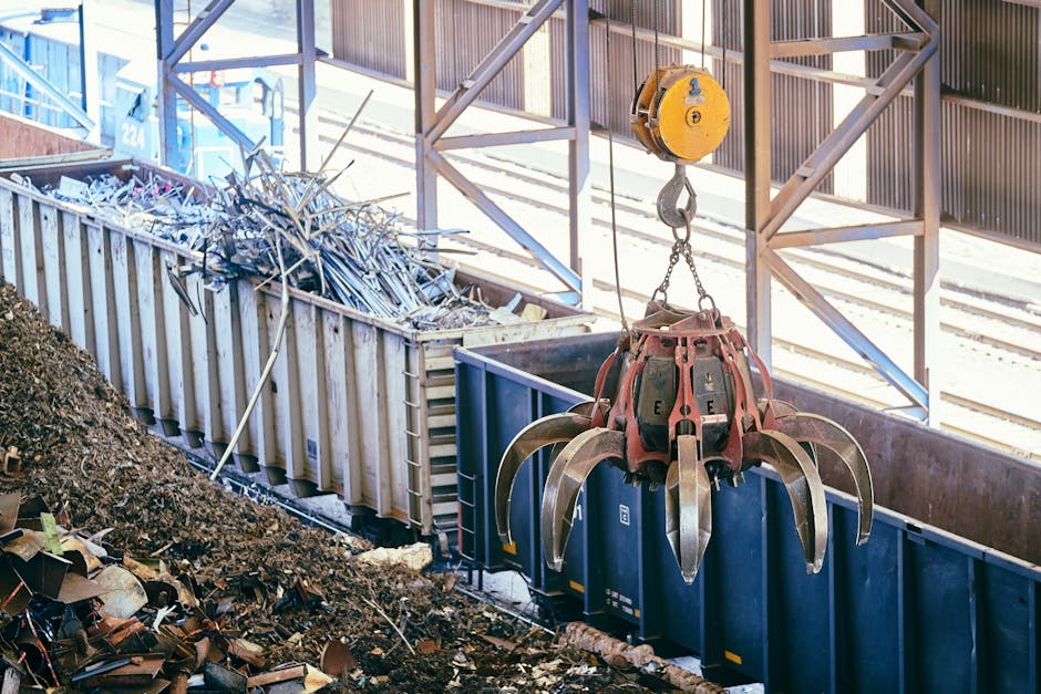A large industrial crane with a mechanical claw extension is hanging above a railway freight yard, lifting a collection of metal scrap and debris. The scene includes a yellow pulley system on a metal 