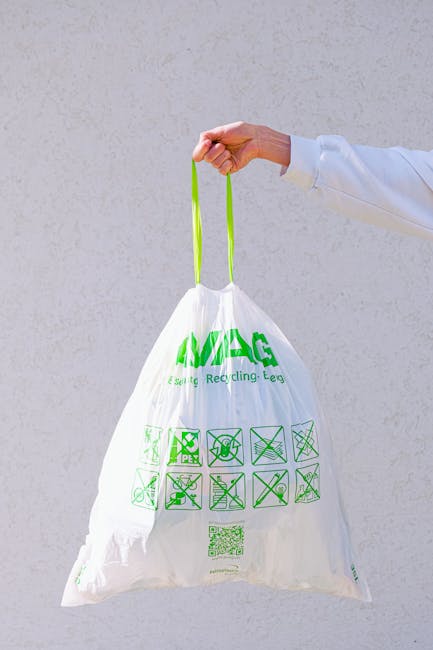 A person's hand, wearing a white sleeve, holding a white recycling bag with green handles and printed recycling symbols against a plain light grey wall. The bag is crumpled and features symbols indica