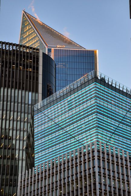The image depicts a modern cityscape with a focus on a cluster of contemporary glass skyscrapers, illuminated by blue and warm yellow lights at dusk. The tallest building features a sleek, angular des