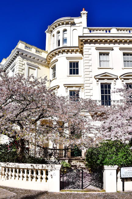 A grand white Victorian-style building with intricate architectural details and large sash windows, set against a vibrant blue sky. In front of the building, several blooming cherry blossom trees with