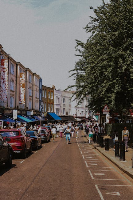 The image depicts a bustling street scene in Notting Hill with a diverse crowd of pedestrians walking along the pavement and crossing the road. On the left side, there are parked cars in various shade