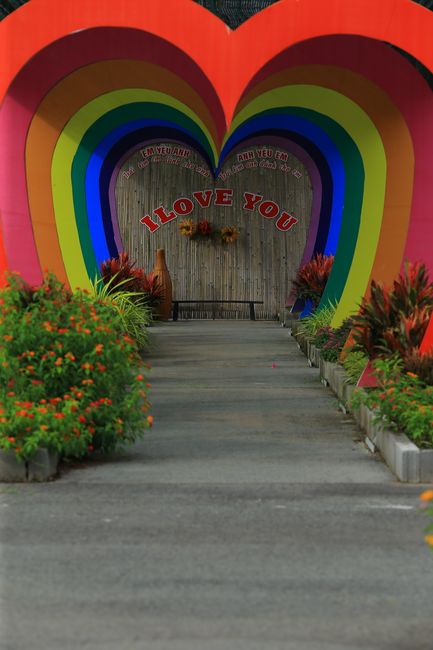 A pathway leading towards a decorative backdrop featuring a large multi-colored rainbow arch painted on a wall, with additional red and pink heart shapes and the words 'I LOVE YOU' displayed prominent