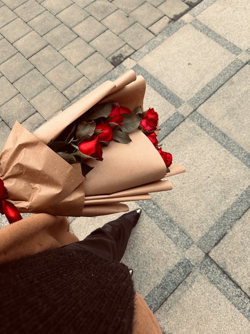 A person holding a bouquet of fresh red roses with dark green leaves, wrapped in light brown kraft paper and tied with a red ribbon. The bouquet is positioned at an angle, showing the vibrant flower h