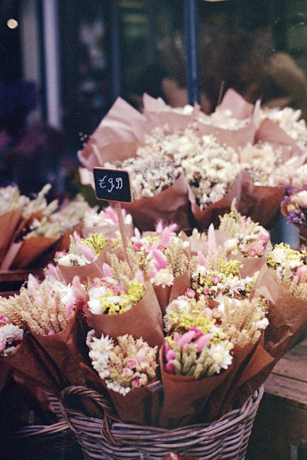 A display of freshly arranged flower bouquets featuring pink tulips, white daisies, and pale yellow blossoms wrapped in light pink and brown craft paper, placed in a woven basket on a wooden surface. 