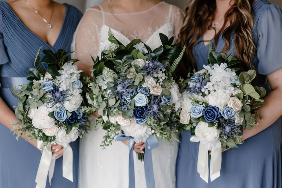 Close-up of three women holding large, round bridal and bridesmaids' bouquets featuring a mix of white and light blue roses, along with white hydrangeas, blue thistle, and green foliage. The bouquets 