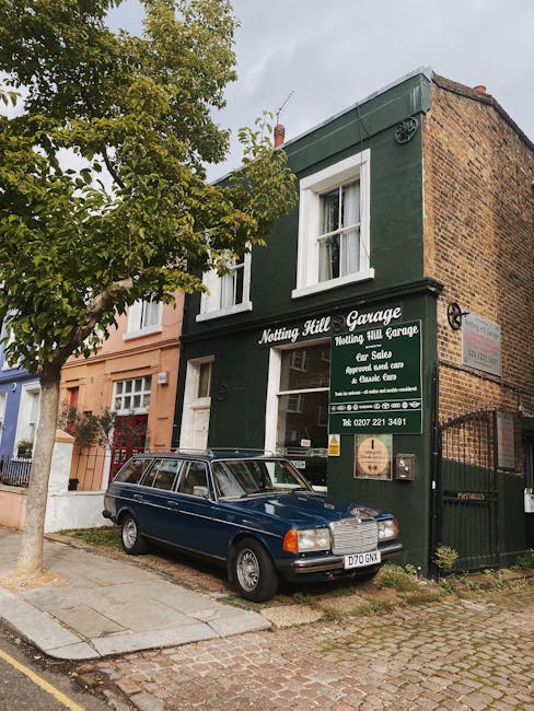 Exterior view of Notting Hill Garage, a small two-storey building painted dark green with white window frames, situated on a cobblestone street. A vintage dark blue station wagon is parked in front, p