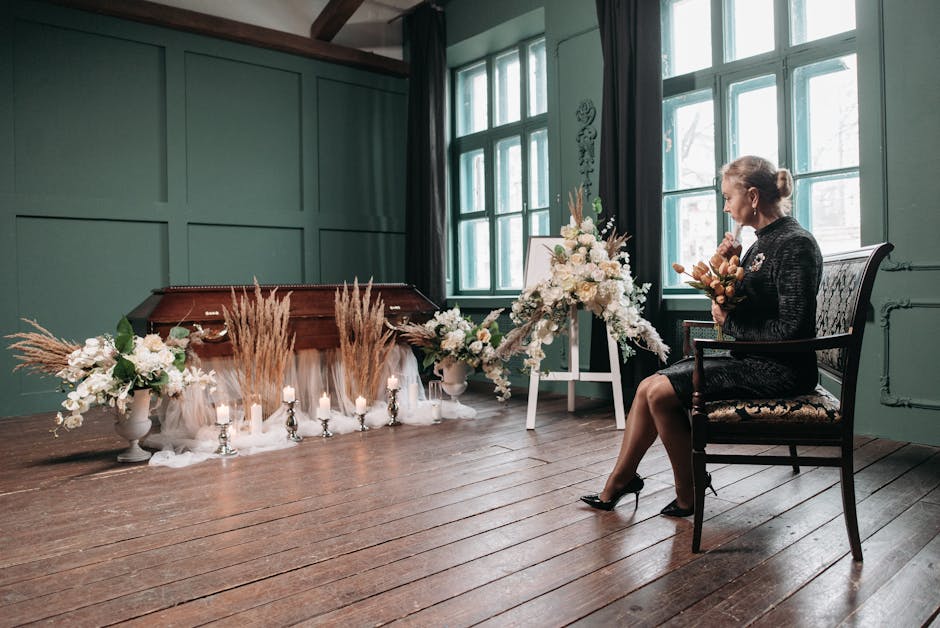 A woman dressed in black, seated on an ornate wooden chair with floral upholstery, is holding a small bouquet of muted-toned flowers and greenery. In front of her, there is a large floral arrangement 