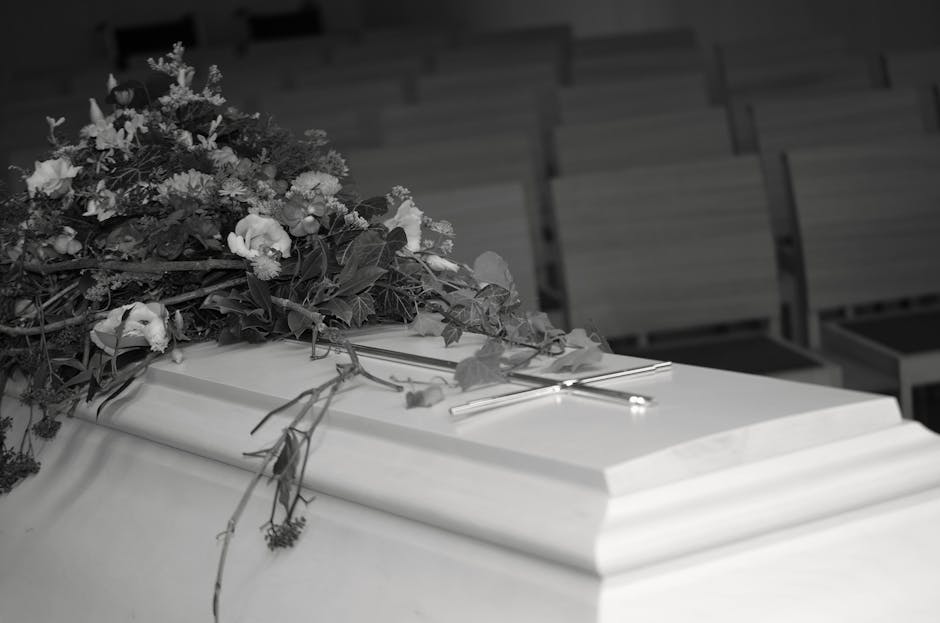 A floral tribute featuring a lush arrangement of fresh flowers, including roses and lisianthus, in soft pastel shades such as white and light pink, arranged atop a white coffin. The bouquet is surroun