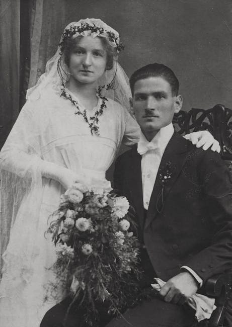 A vintage black-and-white photograph featuring a bride and groom. The bride is dressed in a traditional wedding gown with puffed sleeves and a lace veil adorned with floral embellishments, holding a l